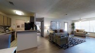 This single-floor residence offers an open-concept layout that connects the main living spaces in a cohesive and functional design. The living area serves as the centerpiece of the home, featuring warm hardwood-style flooring, a textured ceiling, and a comfortable seating arrangement complete with patterned accent chairs, a neutral sofa with colorful throw pillows, and a bold geometric area rug that anchors the space. Framed wall art and a decorative table lamp add refined detail to the room. A pass-through kitchen counter is visible from the living area, providing a convenient bar-style connection to the kitchen, which is equipped with dark cabinetry and a black refrigerator. The kitchen also benefits from its own dedicated lighting, creating a warm, intimate atmosphere. A hallway leads directly from the living area into the bedroom, which is partially visible and features soft, ambient lighting and a neatly arranged bed. Natural light filters in through a window near the entryway, where a tall potted plant adds a fresh, organic element to the space. The overall ambiance is modern and welcoming, with a neutral color palette accented by warm tones throughout.