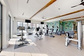 This contemporary fitness center in Chattanooga, TN, is thoughtfully designed to support a full range of workout routines. The space is organized into distinct zones, each tailored to specific training needs. The main cardio area is bright and airy, featuring an impressive row of treadmills, ellipticals, stationary bikes, and a rowing machine, all bathed in abundant natural light streaming through expansive floor-to-ceiling windows that frame outdoor views of the surrounding greenery and development. Exposed wood ceiling beams and a ceiling fan add warmth and character to the otherwise crisp, white interior. A bold, abstract mural in vibrant teal, yellow, and green tones serves as a striking focal point, energizing the space with color and creativity. The strength training zone is well-appointed with a cable machine, power rack, barbells, and a full dumbbell rack, complemented by large mirrors that enhance the sense of openness. A separate studio area features polished concrete flooring, full-length mirrored walls framed by a reclaimed brick accent wall, stability balls, and fitness artwork — ideal for stretching, yoga, or group training. Industrial-style sliding glass barn doors connect the zones, while recessed lighting and mounted flat-screen TVs complete this comprehensive, amenity-rich fitness facility.