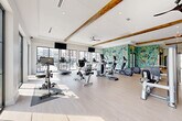 This contemporary fitness center in Chattanooga, TN, is thoughtfully designed to support a full range of workout routines. The space is organized into distinct zones, each tailored to specific training needs. The main cardio area is bright and airy, featuring an impressive row of treadmills, ellipticals, stationary bikes, and a rowing machine, all bathed in abundant natural light streaming through expansive floor-to-ceiling windows that frame outdoor views of the surrounding greenery and development. Exposed wood ceiling beams and a ceiling fan add warmth and character to the otherwise crisp, white interior. A bold, abstract mural in vibrant teal, yellow, and green tones serves as a striking focal point, energizing the space with color and creativity. The strength training zone is well-appointed with a cable machine, power rack, barbells, and a full dumbbell rack, complemented by large mirrors that enhance the sense of openness. A separate studio area features polished concrete flooring, full-length mirrored walls framed by a reclaimed brick accent wall, stability balls, and fitness artwork — ideal for stretching, yoga, or group training. Industrial-style sliding glass barn doors connect the zones, while recessed lighting and mounted flat-screen TVs complete this comprehensive, amenity-rich fitness facility.