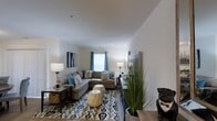 This single-floor residence offers a welcoming and thoughtfully designed living space that combines comfort with modern style. The living area serves as the heart of the home, featuring neutral white walls that create a clean, airy backdrop for the carefully curated furnishings. A generously sized sectional sofa anchors the space, complemented by a patterned area rug in shades of blue, grey, and white that adds visual interest underfoot. Teal accent curtains frame the windows, drawing in natural light while adding a pop of color that ties the room's palette together. The layout is open and functional, with ample room for both relaxing and entertaining. Decorative touches such as table lamps, framed wall art, and tall ornamental grasses lend warmth and character to the space. The smooth, light-toned flooring enhances the sense of openness, while the high ceiling amplifies the spacious feel of the room. Overall, the space strikes a balance between casual comfort and polished, contemporary design, making it an inviting environment for everyday living.