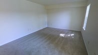 This two-floor space presents a clean, functional layout designed to accommodate a range of needs. The interior is characterized by neutral, light-toned walls that create a bright and open atmosphere throughout. Wall-to-wall carpeting in a muted gray-brown tone runs through the main living areas, offering warmth and comfort underfoot. Natural light filters in through the windows, casting soft reflections across the floor and enhancing the sense of space within each room. The walls feature standard electrical outlets and a wall-mounted vent, indicating practical, well-considered utility placements. The ceiling is smooth with a subtle stepped detail near the upper wall edge, adding a modest architectural element to the otherwise minimalist interior. The overall ambiance is understated and neutral, providing a versatile blank canvas that can easily be adapted to suit personal style or functional requirements. The two-floor configuration allows for a clear separation of living or working areas, contributing to an organized and practical use of the available space. The uncluttered interior and consistent finishes across the space give it a cohesive, move-in-ready feel.
