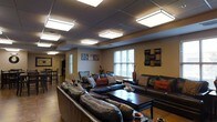 This single-floor communal space is designed to offer a comfortable and inviting environment for gathering and relaxation. The lounge area is anchored by generous dark leather sectional sofas and matching seating arranged around a sleek, dark-toned coffee table, creating an ideal setting for conversation and socializing. Decorative throw pillows in warm earthy tones — including rust, brown, and teal — add a layer of visual interest and comfort to the seating arrangement.

The ceiling features a suspended grid system fitted with multiple large flush-mount light panels, providing bright, even illumination throughout the space. Warm beige and tan walls contribute to the overall welcoming atmosphere, accented by tasteful wall art and decorative pieces that give the space a curated, finished look. A tall decorative vase with branches adds an organic element near the seating area.

Natural light filters in through multiple windows dressed with horizontal blinds, complementing the overhead lighting. In the background, a dining area with dark wood chairs and tables is visible, suggesting a multi-functional layout that supports both leisure and dining. The wood-toned flooring ties the space together, reinforcing a warm and cohesive aesthetic throughout.