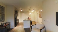This single-floor residence showcases an open-concept design that integrates the kitchen, dining, and living areas into one cohesive space. The kitchen serves as the focal point, anchored by a central island with a granite or stone-look countertop and a built-in sink, providing both functional workspace and casual seating. Sleek dark cabinetry lines the kitchen walls, complementing stainless steel appliances, including a refrigerator and range. Pendant lighting hangs above the island, while a track lighting system illuminates the broader space, creating a warm, well-lit ambiance throughout.

Warm wood-look flooring extends across the entire floor, lending a consistent and inviting character to the space. The open layout allows for a comfortable seating area, as evidenced by the upholstered chair visible in the foreground. Multiple doorways lead to additional rooms, including what appear to be closets or private spaces toward the rear. Neutral wall tones and clean white door and trim details contribute to a modern yet understated aesthetic. The overall atmosphere is welcoming and practical, with thoughtful lighting and finishes that enhance everyday living.