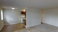 This single-floor residential unit offers a straightforward and functional layout designed for comfortable everyday living. The living area is finished with plush wall-to-wall carpeting, providing a warm and inviting atmosphere, while clean white walls and white baseboards keep the space feeling bright and neutral throughout.

A partial dividing wall separates the living area from the kitchen, creating a sense of definition between the two spaces without fully closing them off from one another. The kitchen features rich, warm-toned wood cabinetry that adds a touch of character to the room. White appliances, including a gas range and oven, complement the cabinetry and contribute to the kitchen's clean, practical aesthetic. The kitchen is finished with tile-style flooring, providing a durable and easy-to-maintain surface that contrasts with the carpeted living area.

The space is illuminated by ceiling-mounted lighting and benefits from a simple, uncluttered design that allows for flexible furniture arrangements. Electrical outlets are conveniently positioned throughout the unit. Overall, this space presents a clean, well-maintained interior with a neutral palette that serves as a versatile foundation for personalization.