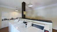 This single-floor laundry facility is designed to serve a shared community, offering a practical and well-organized space for residents. The room is equipped with a row of commercial-grade Speed Queen top-load washers along the front, complemented by a lineup of dryers along the back wall, providing ample capacity for multiple users at once. A dark, wide folding counter runs along the center of the room, supported by a bold structural column finished in dark wood, which serves as a visual anchor in the space. The white walls and light-colored cabinetry contribute to a clean, utilitarian aesthetic throughout. Overhead, industrial-style pendant lights hang from the ceiling, casting even, functional illumination across the room. The machines are arranged in an orderly fashion, maximizing the available floor space while maintaining clear pathways. Informational notices are posted on the walls, ensuring users have access to guidelines and instructions. Overall, the laundry room strikes a balance between functionality and practicality, offering a straightforward, efficient environment suited to everyday use.