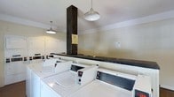 This single-floor laundry facility is designed to serve a shared community, offering a practical and well-organized space for residents. The room is equipped with a row of commercial-grade Speed Queen top-load washers along the front, complemented by a lineup of dryers along the back wall, providing ample capacity for multiple users at once. A dark, wide folding counter runs along the center of the room, supported by a bold structural column finished in dark wood, which serves as a visual anchor in the space. The white walls and light-colored cabinetry contribute to a clean, utilitarian aesthetic throughout. Overhead, industrial-style pendant lights hang from the ceiling, casting even, functional illumination across the room. The machines are arranged in an orderly fashion, maximizing the available floor space while maintaining clear pathways. Informational notices are posted on the walls, ensuring users have access to guidelines and instructions. Overall, the laundry room strikes a balance between functionality and practicality, offering a straightforward, efficient environment suited to everyday use.