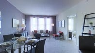This single-floor residence offers a thoughtfully designed layout that balances comfort and contemporary style. The open-plan living and dining area serves as the heart of the space, where soft, neutral carpeting and a muted lavender-gray accent wall create a calm and welcoming ambiance. A bank of large windows dressed with sheer, light-filtering curtains floods the room with natural light, enhancing the sense of openness and airiness. The living area is arranged around a sleek glass coffee table and a plush sofa accented with red and neutral throw pillows, complemented by a bold red accent chair that adds a striking pop of color. A glass-top dining table with dark chairs sits adjacent to the living area, making the transition between spaces seamless and functional. Framed wall art and carefully placed floor and table lamps contribute to the space's polished, curated aesthetic. A hallway visible from the main living area leads to a bedroom, which benefits from its own window, maintaining the consistent theme of natural light throughout. The overall design reflects a modern sensibility with clean lines, a cohesive neutral-and-red color palette, and an efficient use of space across the single floor.