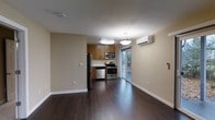 This single-floor space showcases a thoughtfully designed open-concept layout that seamlessly connects the living area and kitchen. The kitchen is equipped with a full suite of stainless steel appliances, including a refrigerator, microwave, and gas range, all complemented by warm wood cabinetry and clean white countertops. Rich, dark hardwood flooring runs throughout the space, adding depth and a sense of warmth to the interior. Two pendant light fixtures hang from the smooth white ceiling, providing focused illumination over the kitchen and adjoining living area. A wall-mounted mini-split air conditioning unit ensures year-round climate control with efficiency. A sliding glass door at the rear of the kitchen floods the space with natural light and provides direct access to an outdoor area, offering a pleasant connection to the surrounding greenery. The walls are finished in a soft neutral tone, creating a clean and versatile backdrop. The open floor plan and thoughtful flow between areas make this a highly functional and inviting single-level space suited for everyday living.
