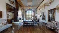 This single-story clubhouse presents a refined, contemporary design throughout its multiple interconnected areas. The main lounge immediately impresses with soaring ceilings, an elegant chandelier, and a dramatic arched window that floods the space with natural light. Dark wood beam accents, wood-look tile flooring, and a sleek double-sided fireplace anchor the room, while curated metallic wall art and plush, neutral-toned seating create a polished yet welcoming ambiance.

A dedicated seating area nearby features tufted accent chairs, coordinating lamps, and a wall-mounted television framed by wood-trimmed windows, offering a comfortable retreat. The game room accommodates a full-sized billiards table beneath pendant lighting, complemented by large windows with wooden blinds.

A café-style bar area provides a curved counter with pendant lights overhead, bar-height tables, and ample seating, alongside a mounted television and bold artwork. The social lounge extends the communal experience with layered seating arrangements, a linear fireplace, and outdoor views through expansive windows.

A polished corridor lined with curated artwork connects the various zones, leading to a formal leasing office featuring a herringbone wood floor, high ceilings with clerestory windows, and a substantial work desk. Throughout, dark wood trim, cohesive artwork, and warm lighting unify the space with a consistently sophisticated character.