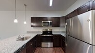 This single-story space offers a well-designed layout with consistent wood-look flooring running throughout, creating a cohesive and polished aesthetic across all rooms. The kitchen serves as a central highlight, outfitted with rich dark espresso cabinetry, granite countertops, and a full suite of stainless steel appliances including a range, built-in microwave, refrigerator, and dishwasher. A peninsula with an integrated sink and pendant lighting above adds both functionality and visual appeal, making it an efficient workspace. The living areas are bright and open, featuring neutral white walls, ceiling fans with integrated lighting, and windows dressed with blinds that allow natural light to filter in. Individual rooms offer generous proportions with clean lines and ample wall space. The bathrooms are finished in a clean, practical style — one features a tub-shower combination with a wall-mounted sink and vanity mirror, while a second bathroom includes a sliding glass-door enclosed shower, dark vanity with granite countertop, and matching wood-look flooring. A wall-mounted mini-split HVAC unit is present in one room, providing supplemental climate control. Overall, the space maintains a clean, modern, and move-in ready character.