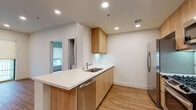 This contemporary single-floor residence is designed with a clean, modern aesthetic throughout. The kitchen serves as a central highlight, featuring warm wood-toned flat-front cabinetry, crisp white quartz countertops, and a full suite of stainless steel appliances including a gas range, built-in microwave, refrigerator, and dishwasher. A peninsula with an integrated sink adds functional counter space while connecting the kitchen to the living area, creating an open, cohesive layout. Recessed lighting illuminates the kitchen and living spaces, complementing the light wood-look flooring that runs throughout the unit. The living area is generously proportioned with clean white walls and sliding glass doors that open to a private balcony, allowing natural light to flow freely into the space. A separate bedroom offers a quiet retreat with a large window fitted with roller shades and warm wood-look flooring. An additional bedroom or flex room provides versatile space with hardwood-style flooring and ample wall space. The bathroom is finished with consistent design elements, including a warm wood vanity with white countertop, a large wall mirror, and matching wood-look flooring, tying together the cohesive and polished character of the entire residence.