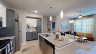 This well-designed single-floor residence offers a cohesive modern aesthetic carried throughout each room. The open-concept kitchen serves as a central feature, showcasing a large granite-topped island with an undermount sink, pendant lighting overhead, and a two-tone cabinet scheme pairing white uppers with slate-gray lowers. Stainless steel appliances, including a refrigerator, range, and built-in microwave, complete the functional workspace. The kitchen flows directly into the living area, which features light wood-style flooring, a ceiling fan, and a glass-panel door providing access to an outdoor balcony and filling the space with natural light.

The unit includes multiple bedrooms, each equipped with ceiling fans, large windows with blinds and draping curtains, and light wood-style flooring. One bedroom features a bold accent wall in deep navy, while another presents a neutral palette. Each bedroom connects to a private bathroom finished with granite-topped vanities, gray cabinetry, and walk-in closets with built-in shelving. The bathrooms also feature soaking tubs and/or shower combinations. A dedicated utility area with laundry hookups adds everyday convenience to this thoughtfully appointed residence.