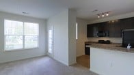 This single-floor space offers an open-concept design that seamlessly connects the living area and kitchen. The living space features neutral-toned carpet, white walls, and large windows that invite generous natural light, including a glass-panel entry door that adds a welcoming brightness to the space. The transition from carpet to hardwood-style flooring marks the entry into the kitchen area, which is equipped with dark espresso cabinetry, granite-style countertops, and black appliances, including a built-in microwave. A kitchen peninsula with a white base provides a functional divide between the two areas, ideal for casual dining or food preparation. Track lighting above the kitchen ensures ample task illumination. The overall layout is clean and functional, with a neutral color palette of whites and grays throughout, creating a cohesive and modern aesthetic. The combination of natural light, open sightlines, and practical design elements makes this a versatile and well-appointed space suitable for everyday living.
