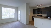 This single-floor space offers an open-concept design that seamlessly connects the living area and kitchen. The living space features neutral-toned carpet, white walls, and large windows that invite generous natural light, including a glass-panel entry door that adds a welcoming brightness to the space. The transition from carpet to hardwood-style flooring marks the entry into the kitchen area, which is equipped with dark espresso cabinetry, granite-style countertops, and black appliances, including a built-in microwave. A kitchen peninsula with a white base provides a functional divide between the two areas, ideal for casual dining or food preparation. Track lighting above the kitchen ensures ample task illumination. The overall layout is clean and functional, with a neutral color palette of whites and grays throughout, creating a cohesive and modern aesthetic. The combination of natural light, open sightlines, and practical design elements makes this a versatile and well-appointed space suitable for everyday living.