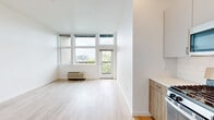 This thoughtfully designed single-floor unit offers a clean, modern aesthetic throughout, characterized by light hardwood flooring, crisp white walls, and an open, airy layout that maximizes the sense of space.

The kitchen is a standout feature, equipped with stainless steel appliances including a gas range, over-the-range microwave, and refrigerator. Cabinetry combines gray upper cabinets with warm wood-toned lower drawers accented by brushed gold hardware, complemented by white quartz countertops and a classic subway tile backsplash. A tall wood-paneled column adds a refined architectural detail to the space.

The living area is bathed in natural light through floor-to-ceiling windows, with direct access to a private balcony offering open outdoor views. The bright, open space flows naturally from the kitchen, making it ideal for both relaxing and entertaining.

The bedroom is generously proportioned, featuring light-toned walls and a wide sliding-door closet that provides ample storage. An additional closet is conveniently located near the entry hallway.

The bathroom continues the warm, contemporary design language with wood-toned cabinetry, an integrated vanity, and light hardwood-style flooring. The unit also includes a wall-mounted utility panel, adding practical functionality. Overall, the space presents a cohesive, well-appointed modern interior with quality finishes throughout.