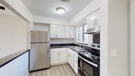 This single-floor unit offers a clean, functional layout with updated finishes throughout. The kitchen is a standout feature, showcasing white shaker-style cabinetry with brushed nickel hardware, dark countertops, and a full suite of stainless steel appliances including a refrigerator, electric range with oven, and a built-in range hood. A single-basin sink sits beneath a window fitted with horizontal blinds, allowing natural light to brighten the space. Light wood-look flooring ties the kitchen together with a fresh, contemporary feel.

The living and sleeping areas are finished with neutral-toned carpet and painted in a soft gray palette, creating a calm, versatile backdrop. Vertical blinds on the windows provide both privacy and light control, while ceiling-mounted light fixtures illuminate each room. The floor plan appears compact yet well-organized, making efficient use of available space.

The bathroom features a tub and shower combination with white subway-style tile, a handheld showerhead, and grab bars for added accessibility. White cabinetry with dark countertops continues the design language from the kitchen, and light wood-look flooring completes the space with a cohesive finish throughout.