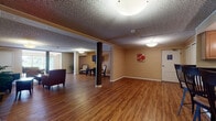 Located at 27825 Detroit Rd in Westlake, Ohio, this single-floor community space offers a welcoming and functional layout designed for gathering and relaxation. The interior features warm hardwood-style flooring that flows consistently throughout, complemented by textured ceilings and neutral earth-tone walls that create a comfortable, inviting atmosphere.

The open-plan common area is anchored by structural columns and divided into several distinct functional zones. A cozy lounge area features a dark upholstered sofa, accent chairs, a wooden coffee table, and coordinating side tables, with wall-mounted artwork adding a decorative touch. A separate seating nook provides wall-mounted flat-screen TV access, furnished with patterned accent chairs ideal for smaller gatherings.

The kitchen area is equipped with light wood cabinetry, dark granite-style countertops, stainless steel appliances, and pendant lighting overhead. A raised breakfast bar accommodates three bar-height chairs, offering a casual dining option. An additional open area near the entrance provides flexible space, currently unfurnished, suitable for a variety of uses.

Flush-mount and pendant light fixtures provide consistent illumination throughout. Multiple doors offer access to additional rooms or exits. The overall design prioritizes communal comfort and versatility, making it well-suited as a shared amenity or multi-purpose community room.