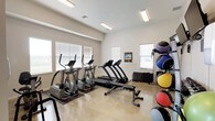 This fitness center is designed to support a comprehensive workout experience within a clean, modern, single-floor layout. The main exercise area features light-toned polished concrete flooring and bright white walls that contribute to an open, energizing atmosphere. Multiple windows fitted with horizontal blinds line the walls, allowing ample natural light to fill the space, complemented by overhead fluorescent lighting.

The cardio zone is well-stocked with several treadmills and elliptical machines positioned along the windowed walls, providing an airy environment for endurance training. A stationary bike adds further variety to the cardio options. Strength training is supported by a multi-functional cable machine, a dedicated weight bench, and a dumbbell rack offering a range of free weights. Stability and functional training equipment includes a rack of medicine balls in assorted sizes and multiple stability balls. Wall-mounted flat-screen televisions are positioned for easy viewing during workouts.

An entry corridor with polished tile flooring connects the main fitness area to restroom facilities, featuring a water fountain and hand towel dispenser for convenience. The hallway leads to clearly marked doorways, keeping the layout organized and accessible. Overall, the space presents a clean, functional, and well-maintained fitness environment suited for a variety of exercise routines.