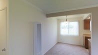 This single-floor space offers a clean, functional layout with a warm and understated aesthetic. The interior features soft, neutral-toned carpet throughout, complemented by crisp white walls and crown molding that lend a polished finishing touch. A textured ceiling adds subtle visual interest overhead, while an exposed structural beam introduces an architectural detail that grounds the space.

Natural light plays a central role in the ambiance, streaming through a large window dressed with vertical blinds, offering views of the greenery outside. A classic pendant light fixture hangs above this window area, providing a focal point that suggests a dedicated dining or gathering space. The interplay of natural and artificial lighting keeps the interior feeling airy and inviting.

A wall-mounted HVAC unit is positioned along one wall, ensuring year-round climate comfort without occupying floor space. The overall design is simple and neutral, making it highly adaptable for a variety of uses. The open, uncluttered layout enhances the sense of spaciousness throughout the interior, creating an environment that is both practical and comfortable.