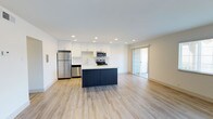 This thoughtfully designed single-floor unit offers a clean, contemporary aesthetic throughout. The heart of the space is an open-concept kitchen and living area, where light wood-look flooring flows seamlessly underfoot. The kitchen is well-appointed with crisp white cabinetry, a patterned tile backsplash, a full suite of stainless steel appliances including a refrigerator, dishwasher, over-the-range microwave, and electric range, plus a central island with a dark navy base and white countertop that provides additional prep and gathering space. A sliding glass door in the living area allows natural light to pour in and offers access to an outdoor balcony with views of surrounding greenery. The spacious living room features large windows fitted with blinds, filling the area with natural light and creating an airy, open ambiance. The bedrooms are carpeted for comfort, with neutral tones and natural light from windows; at least one bedroom includes mirrored sliding closet doors. The bathrooms are finished in a clean, modern style — one featuring a full soaking tub and shower with white subway tile surround, and another with a wide vanity, white countertop, and framed mirror. The overall design is bright, minimal, and move-in ready.