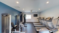 This fitness center offers a clean, functional environment designed to support a full workout routine. The space is finished with a combination of crisp white and deep navy blue accent walls, creating a modern, energizing atmosphere. Recessed lighting throughout the ceiling provides bright, even illumination, while a ceiling-mounted fan ensures comfortable air circulation during workouts. A flat-screen television is mounted on the wall, offering entertainment options for cardio sessions. On one side of the room, a row of treadmills is neatly arranged, providing ample cardio equipment for multiple users simultaneously. On the opposite side, a multi-station cable machine and a weight bench with free weights offer comprehensive strength-training options. The dark rubberized flooring is both practical and durable, providing cushioning underfoot and protecting the equipment. A window along the back wall introduces a degree of natural light into the space. Overall, the fitness center is thoughtfully laid out to maximize usable floor space while keeping circulation paths clear and accessible.