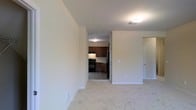 This single-floor residential unit offers a clean, functional layout designed for comfortable everyday living. The living area is finished with plush light-colored carpeting, providing a warm and inviting atmosphere. Neutral-toned walls and flush ceiling lighting create a bright, understated ambiance throughout the space.

An open passageway connects the living area directly to the kitchen, which features rich dark wood cabinetry that extends to the upper and lower sections of the room. The kitchen is equipped with a black range and a stainless steel refrigerator, complemented by tile flooring that contrasts neatly with the carpeted living space. Under-cabinet lighting adds a practical touch to the kitchen's functionality.

A door visible from the living area provides access to additional rooms, suggesting a well-organized floor plan that separates the private and shared areas of the unit. The overall design emphasizes simplicity and practicality, with clean lines and a neutral color palette that allows for versatile interior styling. The single-floor configuration ensures easy accessibility and a cohesive flow between spaces, making this unit a straightforward and efficient place to live.