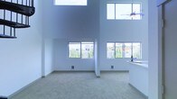 This striking two-story space is defined by its dramatic double-height ceilings, which create an expansive and airy atmosphere throughout the main living area. The open-plan layout is bathed in natural light, courtesy of multiple large windows positioned at both the lower and upper levels, flooding the interior with bright, even illumination throughout the day. Views of surrounding greenery and neighboring structures are visible through the mid-level windows, providing a pleasant connection to the outdoors. The walls are finished in a clean, neutral white, contributing to the space's modern and minimalist aesthetic. Soft, neutral-toned carpeting covers the floor of the main living area, adding warmth and comfort underfoot. A ceiling fan is mounted above, combining form and function in the upper reaches of the space. Adjacent to the main living area, a glimpse of a countertop surface suggests an integrated kitchen or bar area, reinforcing the open and connected nature of the floor plan. The overall ambiance is contemporary and light-filled, with the architectural emphasis on verticality and openness making this a visually impressive and highly functional two-floor space.