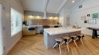 This thoughtfully designed single-floor space offers an open, airy layout that seamlessly blends a fully equipped kitchen, dining area, and lounge into one cohesive environment. Herringbone-patterned hardwood flooring flows throughout, grounding the space with warmth and visual interest.

The kitchen is a standout feature, centered around a concrete-finished island with a white quartz countertop and bar-stool seating. The perimeter cabinetry is finished in natural wood tones, complemented by crisp white counters and a striking metallic penny-tile backsplash. Stainless steel appliances, including a range, microwave, and beverage fridge, make the kitchen both functional and stylish.

The dining area accommodates a large communal table surrounded by mixed seating, positioned beneath a dramatic black-framed glass partition wall that adds an industrial-modern touch and allows natural light to filter through. Tall windows along one wall flood the space with daylight while offering views to the outdoors.

The lounge area is furnished with plush green accent chairs and a round coffee table, creating an inviting nook for relaxed conversation. A wall-mounted TV and shuffleboard table add a recreational dimension to the space. Decorative hexagonal wall planters filled with air plants add a distinctive organic accent. Vaulted, beam-detailed ceilings lend the entire space a sense of generous height and openness.