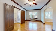 This single-floor unit showcases a blend of classic craftsman architectural details and timeless design elements. Rich, dark-stained wood trim frames doorways, baseboards, and crown moldings throughout, creating a cohesive and characterful interior. Warm hardwood floors extend across the main living areas, reflecting natural light that streams in through double-hung windows framed in matching dark wood.

The living area is highlighted by an elegant arched doorway that connects rooms, adding architectural charm and an open sense of flow. Textured plaster walls lend additional character to the space. A ceiling fan with a light fixture serves the bedroom, which also features a cast-iron radiator beneath the window, a nod to the building's vintage origins. A closet with hanging space adds practical storage.

The kitchen retains its classic appeal with white cabinetry featuring glass-panel doors, a black-and-white diamond-patterned tile floor, and built-in shelving in an adjoining pantry area. The bathroom continues the vintage aesthetic with a classic hexagonal tile floor adorned with a blue floral pattern, a soaking tub, and a radiator. A rich wood door with ornate detailing completes the space, reinforcing the unit's well-preserved, period-appropriate character throughout.