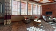 This thoughtfully designed single-floor common area offers a warm and inviting atmosphere through a combination of classic architectural details and carefully curated furnishings. Large, multi-pane windows fitted with warm wood-toned blinds and decorative patterned curtains line the walls, flooding the space with generous natural light throughout the day.

The main lounge area is anchored by a rich, wood-paneled entertainment wall housing a flat-screen television, surrounded by plush sofas and upholstered armchairs accented with bold red and patterned throw pillows. Gold-framed glass accent tables add a refined touch, while patterned carpeting in muted earth tones grounds the seating arrangements. Coffered ceiling details with warm wood trim lend an air of architectural sophistication.

A dedicated dining nook features a round marble-top table encircled by striking red leather chairs with nailhead trim, positioned beside tall windows for a bright and cheerful ambiance. Adjacent to this area, a compact bar counter with white upholstered barstools provides an additional casual seating option.

The kitchenette is equipped with gray shaker-style cabinetry, dark granite countertops, stainless steel appliances including a full-size refrigerator and built-in microwave, and hardwood flooring — offering functional convenience within the elegantly appointed space.