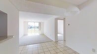 This single-floor space offers a clean, open layout filled with abundant natural light. The main living area showcases a seamless transition between light-tiled flooring in the entry zone and plush carpeting in the living space beyond, clearly delineating functional areas without the need for walls. A textured ceiling adds subtle dimension overhead, while crisp white walls throughout create a bright, airy atmosphere.

The focal point of the living area is a large sliding glass door that opens to a balcony with a railing, framing views of lush greenery and neighboring structures. This feature draws natural light deep into the interior, enhancing the sense of spaciousness. The open-plan design allows for flexible furniture arrangements to suit a variety of living styles.

A doorway visible toward the rear of the space leads to an additional room, suggesting a practical and connected floor plan. The overall aesthetic is minimal and neutral, providing a versatile blank canvas. The combination of light flooring, white surfaces, and generous natural illumination gives the space a welcoming, well-lit character throughout.