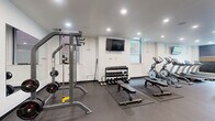 This fitness center at 74 Franklin St in Buffalo, NY, offers a spacious and thoroughly equipped workout facility spread across a single floor. The space is designed with functionality and motivation in mind, featuring durable rubber flooring throughout to support a wide variety of exercises and equipment. Recessed lighting illuminates the entire area evenly, while several windows allow natural light to filter in, creating a bright and energizing atmosphere.

The cardio zone is well-stocked with multiple treadmills, elliptical machines, and stationary cycling bikes, each positioned to provide ample space for users. Wall-mounted flat-screen televisions are strategically placed throughout the facility to keep members engaged during workouts. The strength training area includes a multi-station cable machine by True Fitness, a weight bench, a full rack of dumbbells in graduated sizes, and a barbell setup with weight plates.

A dedicated cycling corner features spin bikes positioned in front of a bold, eye-catching accent wall adorned with a hexagonal graphic design and the motivational phrase "Work Sweat Achieve." Full-length mirrors along one wall enhance the sense of space and allow users to monitor their form. Stability balls and additional functional fitness accessories round out the available equipment, making this a comprehensive facility suited for a wide range of fitness goals.