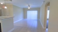 This single-floor space offers a clean, functional layout with a welcoming and well-lit ambiance throughout. The main living area is finished with light-toned ceramic tile flooring that extends seamlessly across the room, contributing to a sense of openness and easy maintenance. Crown molding lines the ceiling, adding a subtle architectural detail that elevates the overall finish of the space.

A ceiling fan with an integrated light fixture is centrally positioned, providing both air circulation and ambient lighting. The room is bathed in natural light courtesy of a large sliding glass door fitted with vertical blinds, offering both brightness and privacy control. The door serves as a focal point of the living area and hints at outdoor access beyond.

The walls are painted in a soft, neutral tone that complements the light flooring and creates a calm, airy atmosphere. White baseboards frame the perimeter of the room, adding a crisp contrast to the warm wall color. A partial view of an adjacent area — likely a kitchen or dining space — is visible, suggesting a connected, open-concept flow between living zones.

Overall, this space presents a straightforward and practical interior characterized by consistent finishes, ample natural light, and a layout designed for comfortable everyday living.