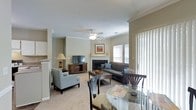 This single-floor space offers an open-plan design that seamlessly integrates the living and dining areas into one cohesive environment. The living area is anchored by a tiled fireplace with a classic mantle, above which framed artwork adds a decorative focal point. A dark flat-screen TV sits on a wooden media console nearby, while a ceiling fan with integrated lighting fixture illuminates the space from above. Crown molding runs along the perimeter of the ceiling, lending a refined architectural detail throughout. The room is finished with plush wall-to-wall carpeting in a neutral tone, contributing to the space's warm and comfortable ambiance. Seating includes a dark sofa accented with blue pillows and a pair of upholstered armchairs in soft grey, arranged around a wooden coffee table. The dining area, positioned toward the front of the space, features a round glass-top table with dark wooden chairs. Large windows dressed with vertical blinds allow natural light to filter in generously, creating a bright and airy atmosphere. Overall, the space presents a traditional yet functional layout suited for everyday living and entertaining.