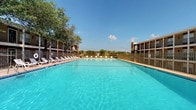 This outdoor pool area offers a well-appointed retreat designed for relaxation and recreation. The centerpiece is a generously sized swimming pool filled with clear, turquoise water, complemented by a separate spa or hot tub situated along the pool deck. The surrounding concrete deck provides ample space for lounging, with multiple chaise lounge chairs and seating arranged to take full advantage of the open sky and sunny exposure.

The pool area is enclosed by a wrought iron security fence, providing both safety and a defined boundary for the space. Mature trees line portions of the perimeter, offering natural shade and a touch of greenery against the open sky. Multi-story residential buildings frame the background, providing a sense of community context.

A dedicated pool house or amenity building is accessible via a rustic wooden-framed entryway, featuring stucco exterior walls and a wall-mounted light fixture. Large glass sliding doors open directly onto the pool deck, allowing for a seamless indoor-outdoor connection and clear sightlines to the water. Posted pool rules and signage are visible near the entry, ensuring guests are informed of guidelines. The overall atmosphere is bright, open, and inviting, making this pool area a standout communal amenity suited for warm-weather enjoyment.