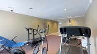 This dedicated fitness space offers a well-equipped environment designed for convenient at-home exercise. The room is finished with warm, neutral-toned walls and dark carpeting that provides cushioning underfoot, creating a practical and comfortable workout setting. Recessed lighting illuminates the space evenly, complemented by a textured ceiling that adds a traditional aesthetic. The layout accommodates multiple pieces of cardio equipment, including at least two treadmills and an elliptical trainer, along with what appears to be a cable pulley machine, allowing for a varied range of workouts. Crown molding along the upper perimeter of the walls adds a refined touch to the otherwise utilitarian space. The room provides ample floor space between machines, ensuring safe and comfortable movement during exercise. Ventilation is supported by visible ceiling vents, contributing to air circulation during workouts. Overall, this fitness room is thoughtfully arranged to support a consistent exercise routine, combining functional equipment placement with clean, straightforward finishes that keep the focus on performance and utility.