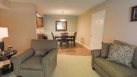 This single-floor space presents an open, functional layout that seamlessly connects the living room, dining area, and kitchen. The living room is carpeted and furnished with comfortable seating, creating a relaxed and inviting atmosphere. Warm neutral tones on the walls contribute to an overall sense of calm throughout the space.

Transitioning from the living area, the flooring shifts to warm-toned hardwood as it flows into the dining space. A dark wood dining table with matching chairs serves as the centerpiece of this area, anchored beneath a pendant light fixture that provides focused overhead lighting. A framed mirror on the accent wall adds visual depth and reflects natural light from a nearby window, brightening the room.

Adjacent to the dining area, the kitchen is partially visible and features wooden cabinetry, offering practical storage solutions. The open sightlines between the kitchen, dining, and living spaces make the layout feel cohesive and well-connected, ideal for everyday living and casual entertaining. The overall design is straightforward and unpretentious, prioritizing comfort and functionality.