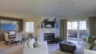 This two-floor space offers a welcoming living area characterized by a clean, neutral color palette of soft beige and white tones. The living room serves as a central gathering space, anchored by a white-tiled fireplace with a classic black insert, above which a flat-screen television is mounted. Decorative metallic leaf wall art flanks the TV, adding an organic, artistic touch. Blue glass vases and accent pieces provide subtle pops of color against the light walls. Natural light filters in through multiple windows, including a sliding glass door that opens to an outdoor balcony, where additional seating is visible, extending the living space outdoors. The room is furnished with a plush white sofa and complementary wicker coffee tables, creating a relaxed yet refined atmosphere. Lush green plants are incorporated throughout, adding life and texture to the space. Neutral-toned carpet underfoot contributes to the overall warmth and comfort of the interior. The overall ambiance is bright, airy, and casually elegant, well-suited for both everyday living and entertaining.