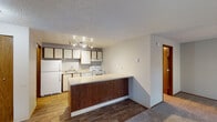 This single-floor residence offers a practical and functional layout designed for comfortable everyday living. The kitchen is a standout feature, equipped with white appliances including a refrigerator, electric range, and dishwasher, complemented by light-toned countertops and a mix of white upper cabinetry with warm wood-toned lower cabinets. A track lighting system illuminates the workspace, while a peninsula breakfast bar with matching wood-panel detailing connects the kitchen to the adjacent living area, creating an open and cohesive flow between spaces. The flooring transitions from wood-look planks in the kitchen and entryway to plush wall-to-wall carpeting in the living room, adding warmth underfoot. The living area is generously proportioned, featuring a large sliding glass door with vertical blinds that allow abundant natural light and provide views of the surrounding outdoor greenery. A baseboard heating system lines the perimeter, ensuring consistent warmth throughout. The bathroom is neatly finished with a wood-framed mirror, a three-bulb vanity light fixture, a single-basin sink set in a laminate countertop with wood-toned cabinetry below, and wood-look flooring. A textured ceiling is present throughout, adding a classic residential character to the overall space.