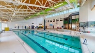 Located at 25 Woodridge Cres in Ottawa's west end, this indoor aquatic facility spans nearly 4,900 square feet on a single level, offering a comprehensive range of aquatic amenities in a bright, well-designed environment. The centerpiece is a multi-lane lap pool clearly marked with lane dividers, suited for both recreational swimming and structured fitness. Adjacent to the lap pool is a shallower leisure pool area featuring a distinctive umbrella-style spray feature, making it ideal for family and instructional use. A spacious octagonal hot tub with decorative mosaic tile detailing provides a hydrotherapy area. The architecture is a defining feature of the facility: soaring vaulted ceilings incorporate warm natural wood planking supported by white structural trusses accented with bold green steel beams, creating an open, airy atmosphere. Expansive clerestory windows and large ground-level windows along the perimeter flood the pool deck with natural light and offer views of the surrounding greenery. The entrance area features a dramatic floor-to-ceiling glass curtain wall. Throughout, non-slip mosaic tile surfaces line the pool deck and surrounding walkways, with green and white patterned tile borders providing visual detail. The facility also includes accessible pool entry equipment, safety signage, and a glazed corridor connecting to additional amenities.