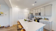 Located at 4086 Silverside Way in Johns Island, SC, this two-story home offers a thoughtfully designed layout across two floors. The main level features an open-concept living, dining, and kitchen area bathed in natural light from multiple windows. The kitchen is a standout, boasting a large quartz-topped island with bar seating, gray shaker-style cabinetry, a white subway tile backsplash, and a full suite of stainless steel appliances. Light wood-tone flooring flows seamlessly throughout the main level, connecting the kitchen to the bright dining area and into the spacious living room, which features vaulted ceilings and a ceiling fan. The upper floor houses three bedrooms, including a generously sized primary bedroom with an en-suite bathroom, alongside two additional well-lit bedrooms and two more full bathrooms. Each bathroom is clean and functional, with one featuring a tub-shower combination with gold-tone fixtures. Hallways connect the upstairs rooms, with a staircase providing easy access between floors. The home's consistent white walls, recessed lighting, and wood-look flooring lend a cohesive, modern aesthetic throughout, making this Johns Island property a comfortable and practical residence.