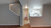This two-story residence offers a well-organized layout designed for comfortable everyday living. The main level greets you with warm hardwood-style flooring that extends throughout the open living area, creating a cohesive and inviting atmosphere. A staircase with white-painted railings and trim rises gracefully along one wall, providing access to the upper floor while adding a clean architectural element to the space.

The kitchen is positioned adjacent to the living area and features a raised peninsula counter that serves as a natural divider between the two spaces. White cabinetry lines the walls, complemented by white appliances including a refrigerator and a range, all set beneath under-cabinet lighting that brightens the workspace. The countertops offer ample prep space for everyday cooking needs.

A central hallway connects the main living area to additional rooms at the rear of the floor, with carpeted spaces visible beyond the corridor, suggesting a comfortable bedroom or secondary living area. Neutral gray walls and white trim work throughout the main level lend a fresh, clean aesthetic. The home's two-floor configuration allows for a natural separation between living and private spaces, making it a practical and functional layout overall.