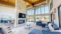 This generously sized single-floor community clubhouse is defined by its dramatic double-height ceilings clad in warm wood paneling, supported by exposed structural beams that lend an inviting, modern-rustic character. The open-plan layout flows seamlessly across light-toned hardwood-style flooring, connecting distinct functional zones within one expansive volume.

At the heart of the space stands a floor-to-ceiling natural stone fireplace, anchored by a wall-mounted television, serving as a striking architectural focal point. Adjacent to it, a comfortable lounge area features plush sectional seating arranged around a round coffee table, ideal for relaxed gatherings. Overhead, a statement chandelier adds an elegant touch.

Large-format, floor-to-ceiling black-framed windows wrap multiple walls of the space, flooding the interior with abundant natural light and providing panoramic views of the surrounding outdoor landscape. A dedicated dining area accommodates a long communal table with upholstered chairs, complemented by bar-height counter seating along the perimeter windows. A well-appointed kitchenette area with vibrant teal cabinetry adds a lively accent.

A recreation zone with a foosball table and casual lounge seating rounds out the space, making this clubhouse a well-balanced, multi-functional gathering destination suited for socializing, dining, and leisure.