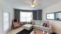 The Montreal is a well-appointed 2-bedroom, 2-bathroom residence that combines modern design with everyday comfort across a single floor. The living area serves as the heart of the home, featuring neutral-toned walls accented by a bold charcoal feature wall that lends a sophisticated, contemporary feel. A ceiling fan with integrated lighting provides both comfort and ambiance overhead, while large windows dressed with white sheer curtains and blinds allow natural light to fill the room. The space accommodates a generous sectional sofa paired with a patterned accent chair, creating an inviting and versatile seating arrangement centered around a rich, warm-toned coffee table. Plush area rugs define the living zone and add layers of texture to the carpeted flooring. The overall design aesthetic is modern and understated, with a neutral color palette punctuated by warm accent tones throughout. The single-level layout ensures easy flow between living spaces, making the residence both practical and welcoming. The Montreal offers a thoughtfully designed home that balances style and functionality for comfortable everyday living.