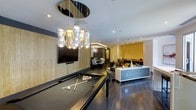 This single-floor space is designed as a sophisticated entertainment and social hub, combining multiple functional zones within an open, flowing layout. The room is anchored by a full-size pool table positioned beneath a striking cluster pendant chandelier, composed of individually shaped glass bulbs suspended from a circular mirrored ceiling medallion — serving as the visual centerpiece of the space.

Dark hardwood flooring runs throughout, providing a sleek, cohesive foundation that contrasts with the light-toned wall panels. Recessed ceiling lighting supplements the statement fixture, ensuring the space is well-lit across all zones.

Beyond the billiards area, a lounge seating arrangement with a sofa and accent cushions in warm tones offers a relaxed gathering spot, complemented by a teal-finished sideboard. A wall-mounted flat-screen television with a soundbar is integrated into the adjacent wall panel.

Further into the space, a dining area with a table and chairs connects to what appears to be a bar or refreshment station, framed by a bold mustard-yellow accent wall with decorative geometric detailing. The overall ambiance is modern and upscale, with carefully layered design elements throughout.