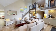 Located at 4901 Tall Timber Dr in Raleigh, NC, this single-floor space makes a striking architectural impression with its dramatic double-height ceilings in the main living area. A sleek metal-and-cable staircase leads to an open mezzanine level overhead, adding vertical depth and a contemporary industrial touch to the overall design.

The main living area is generously proportioned, anchored by neutral-toned sectional sofas arranged around a dark wood coffee table atop a patterned area rug. Colorful framed artwork adorns the tall walls, while an adjacent seating area features a stone-surround fireplace with a wall-mounted television above it, providing a warm and inviting focal point.

The kitchen is appointed with dark cabinetry, light quartz countertops, and textured stone tile backsplash walls that add rich visual interest. Recessed lighting illuminates the workspace, while a large window above the sink frames views of an outdoor deck and lush greenery beyond.

A bright sunroom-style nook lined with oversized windows on two walls invites abundant natural light, offering a tranquil retreat with comfortable lounge seating. Throughout the space, warm hardwood-style flooring flows continuously, unifying each area with a cohesive and welcoming aesthetic. The overall ambiance is modern yet comfortable, balancing open, airy volumes with intimate, well-defined spaces.