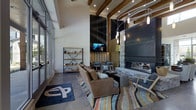 This single-floor lounge space makes a striking first impression with its soaring double-height ceilings supported by bold exposed wood beams, creating an open and airy atmosphere. The interior design blends rustic and contemporary elements seamlessly, anchored by a commanding stone fireplace that serves as the room's focal point. Adjacent to the fireplace, a large mounted television and a dramatic vertical slatted wood accent wall add depth and visual interest to the space.

The seating area is thoughtfully arranged with plush leather-toned sofas and modern accent chairs, centered over a vibrant multi-striped area rug that introduces warm pops of color against the polished concrete floors. A decorative open-frame shelving unit offers both functional storage and a curated display area. Overhead, a sculptural pendant light fixture adds an artistic touch, complementing the recessed lighting throughout the ceiling.

Floor-to-ceiling windows along one wall flood the interior with abundant natural light, enhancing the welcoming and social ambiance of the space. The overall aesthetic is an inviting blend of industrial, modern, and lodge-inspired design — making this lounge an ideal gathering space for relaxation and community engagement.