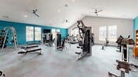 Located at 5001 Willowgate Loop in Fort Myers, Florida, this fitness center offers a comprehensive workout environment across a generously sized single-floor layout. The space is divided into two distinct areas: a main gym floor and a dedicated spin studio, each thoughtfully designed to serve different exercise needs.

The main gym area features soaring vaulted ceilings that create an open, airy atmosphere, accentuated by bold teal accent walls and crisp white upper walls. Multiple ceiling fans ensure comfortable airflow throughout. Natural light streams in through numerous windows along the perimeter, providing views of the surrounding community. The floor is finished with durable, multi-toned carpet tiles. Equipment includes a wide array of strength machines, cable systems, free weights with a dedicated rack, weight benches, treadmills, ellipticals, and a stationary bike. Floor-to-ceiling mirrors line portions of the walls, enhancing the sense of space. A wall-mounted TV is centrally positioned for entertainment during workouts.

The separate spin studio offers a quieter, focused environment with its own ceiling fan, recessed lighting, and large windows. A full-length mirror spans one wall, and the room accommodates multiple cycling bikes on coordinating carpet tile flooring. The studio connects directly to the main gym via an interior door, providing seamless access between both areas.