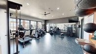 Located at 704 Dale Ln in White Settlement, TX, this well-equipped fitness center offers a comprehensive workout environment within a single, open-plan exercise room. The space features durable slate-blue flooring throughout and is illuminated by a combination of recessed lighting and a ceiling fan, maintaining a comfortable atmosphere during workouts.

Large, dark-framed windows line one wall, flooding the room with natural light and providing views of the surrounding outdoor area. The cardio zone includes a treadmill, elliptical trainers, a rowing machine, and a stationary bike, all positioned to take advantage of the natural light. On the opposite side, a full-length mirror wall visually expands the space and supports strength training, which is further facilitated by a rack of dumbbells, a cable machine, a weight bench, and a selectorized strength machine.

A vibrant orange accent wall serves as a focal point near the entrance, displaying medicine balls and stability balls on wall-mounted holders alongside a water fountain and posted fitness center policies. A wall-mounted flat-screen TV adds a multimedia element to the strength training area. The space also benefits from a dedicated cable functional trainer, rounding out the full-body workout options available. The overall design is modern, clean, and functional, catering to a wide range of fitness needs.