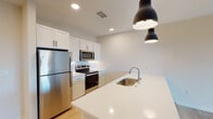 This single-floor residence is designed with a clean, contemporary aesthetic that emphasizes open living and functional design. The kitchen serves as a central hub of the layout, featuring white shaker-style cabinetry, stainless steel appliances including a refrigerator, electric range, and microwave, and a large island with a white countertop and undermount sink. Matte black pendant lights hang above the island, adding a modern contrast to the bright, neutral palette. Recessed lighting complements the pendants throughout the kitchen and living areas.

Light hardwood-style flooring extends through the kitchen and main living spaces, contributing to a cohesive, airy feel. The open living area offers generous wall space and connects seamlessly to the kitchen, making it well-suited for flexible furniture arrangements.

The bedrooms feature soft carpeting and neutral walls, with at least one room benefiting from a window with natural light and outdoor greenery views. The bathroom is finished with a white vanity, light tile flooring, and a glass-enclosed shower stall, maintaining the unit's consistent modern and clean design throughout.